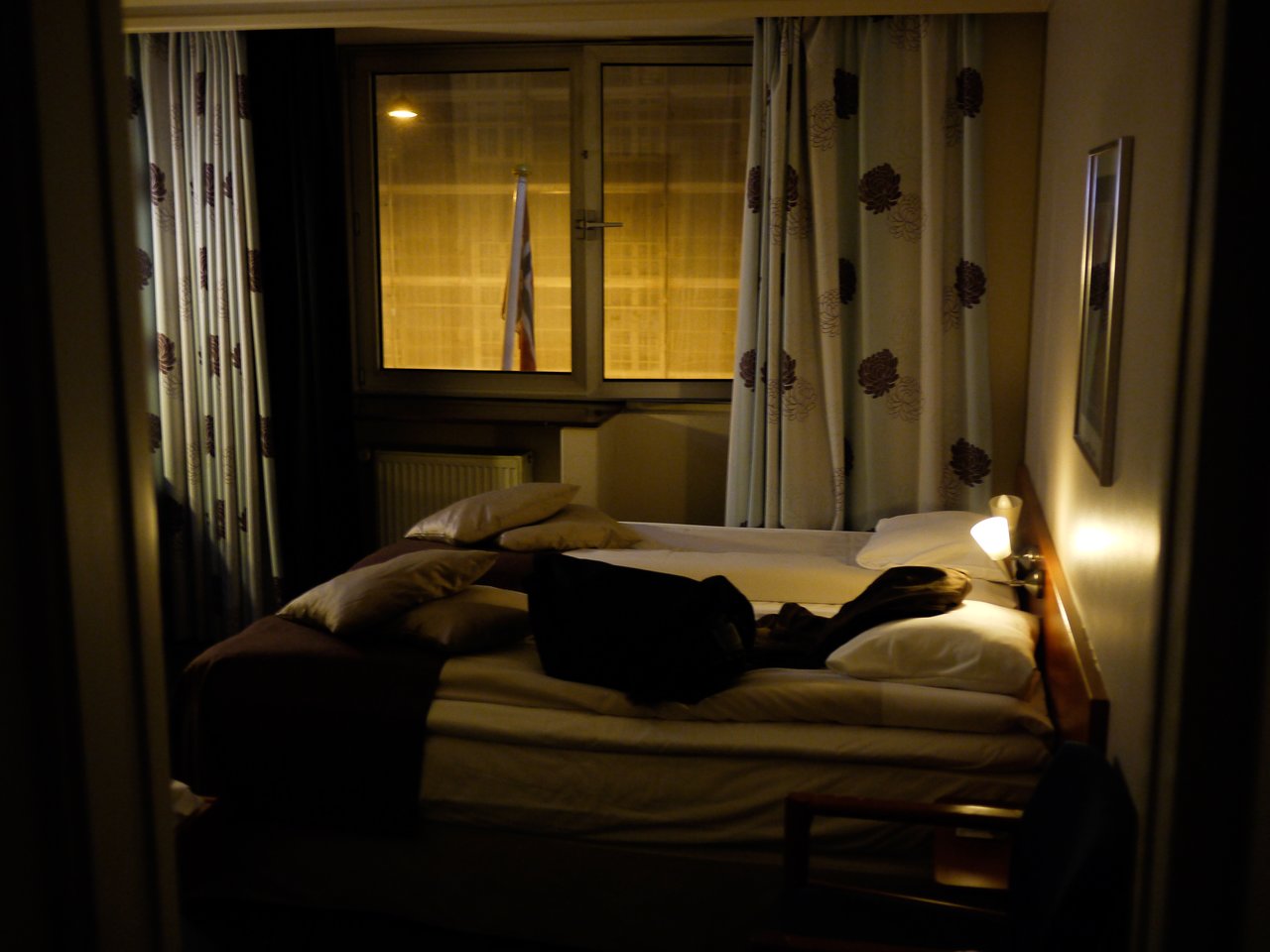 A dimly lit hotel room with an unmade bed, scattered pillows, and a bag resting on top.