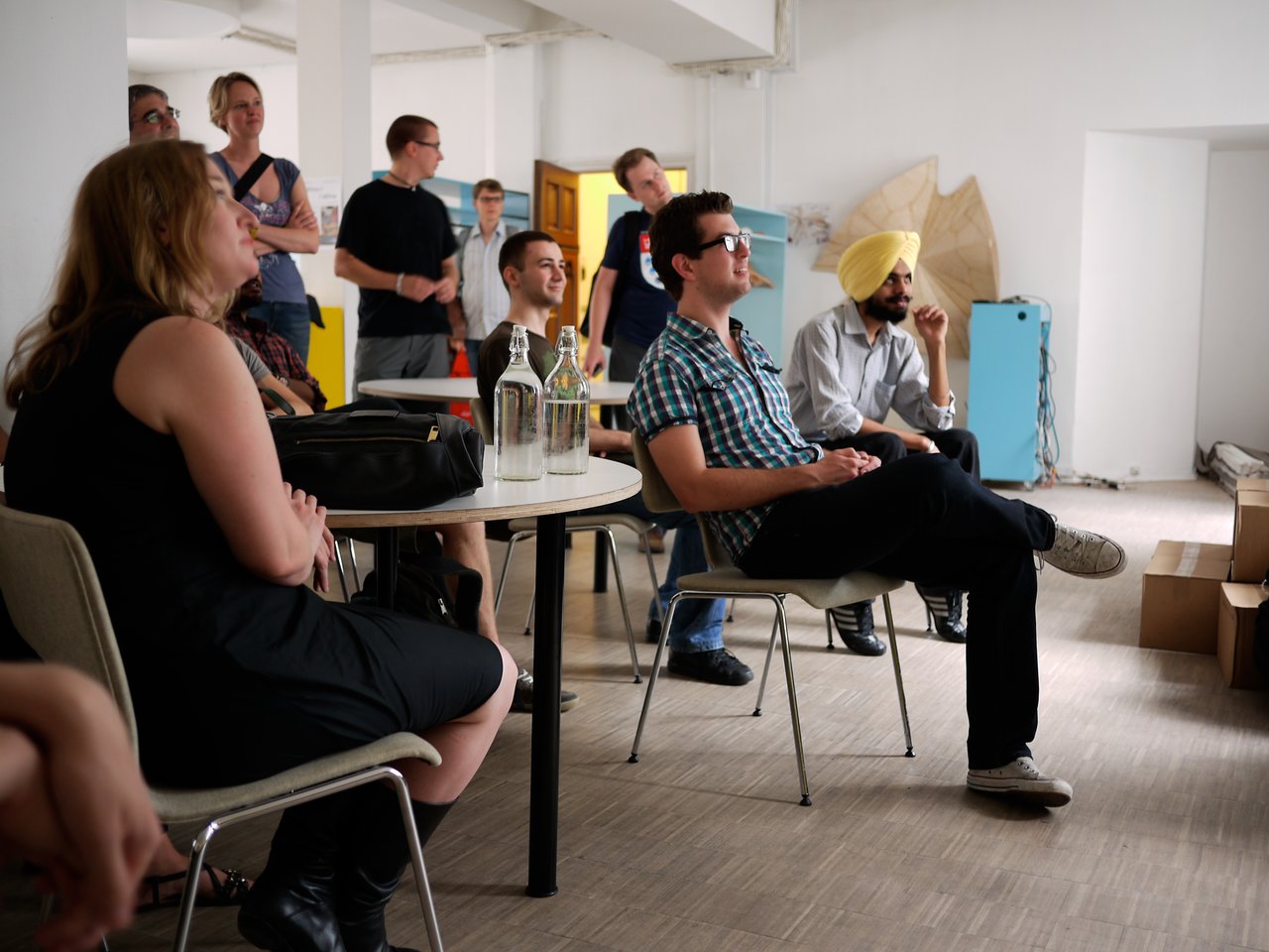 A group of people sits and stands in a room, attentively watching a presentation or discussion.