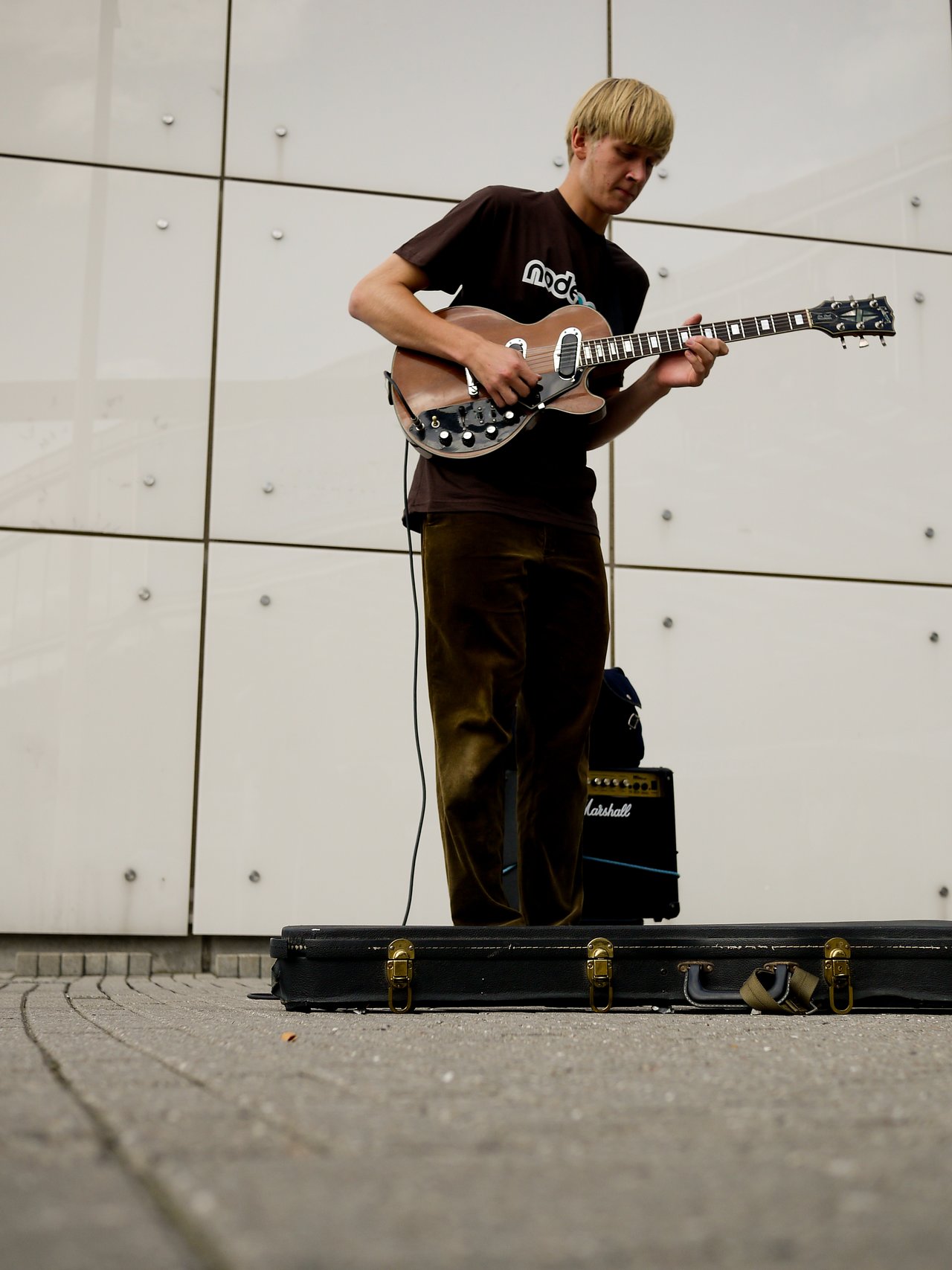 A musician plays an electric guitar outdoors, with an amplifier nearby and an open guitar case on the ground.