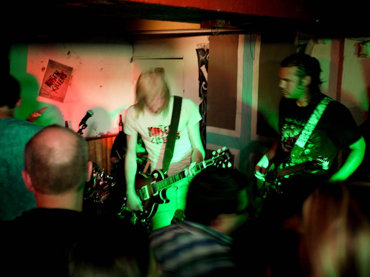 A live band performs in a dimly lit venue, with two guitarists playing energetically as the crowd watches.