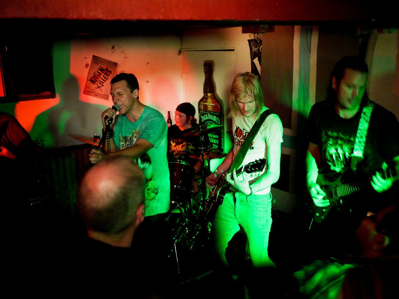 A band performs live in a dimly lit venue, with a singer and two guitarists playing energetically for the crowd.