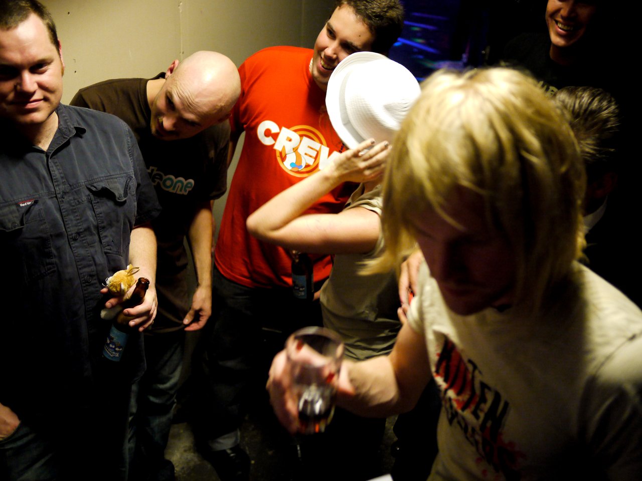A group of people socializing backstage, some holding drinks and food, with one person wearing a blonde wig.