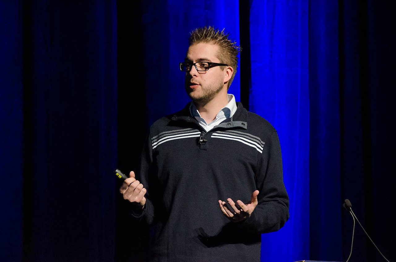 A speaker on stage at DrupalCon Denver 2012, delivering a keynote presentation while holding a remote control.