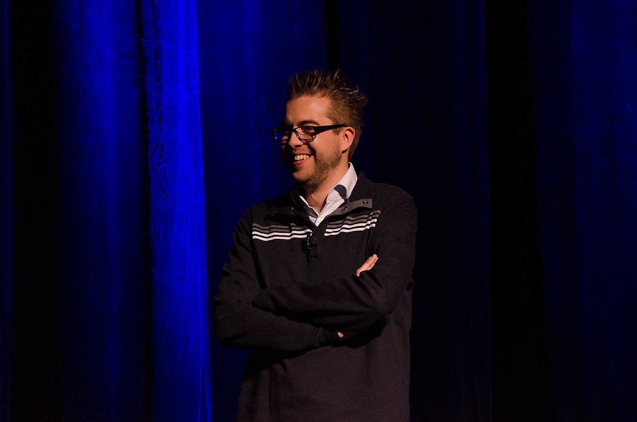 A speaker stands on stage with arms crossed, smiling during the DrupalCon Denver 2012 keynote.
