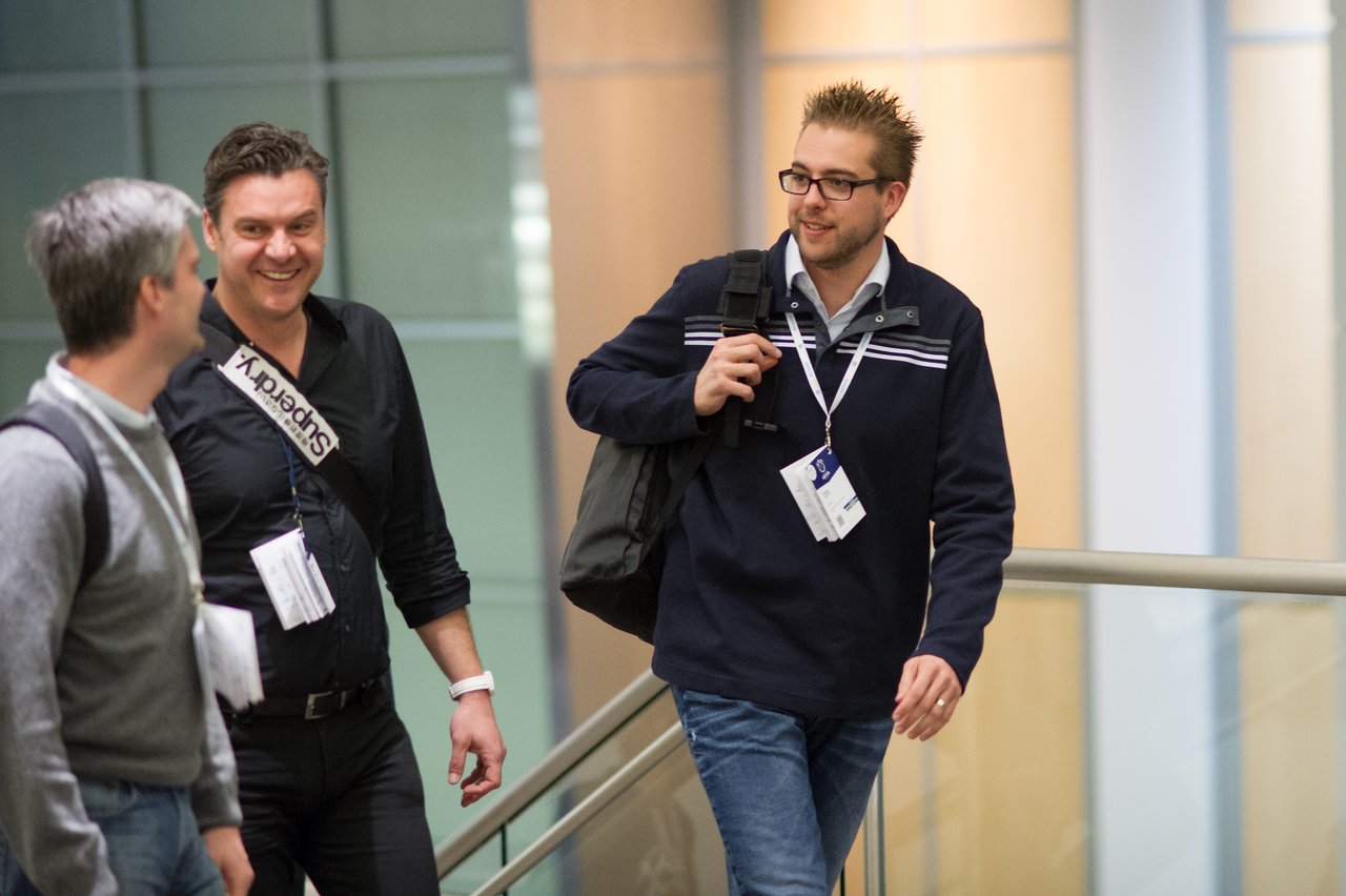 Three conference attendees walk together indoors, wearing name badges and carrying bags, engaged in conversation and smiling.