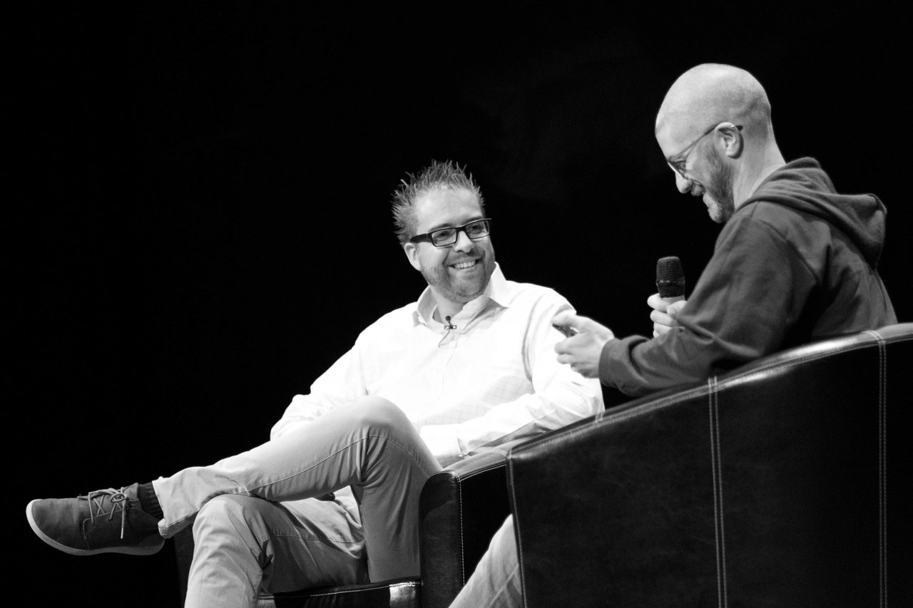 Two people sit on stage in a discussion, one holding a microphone and the other smiling.