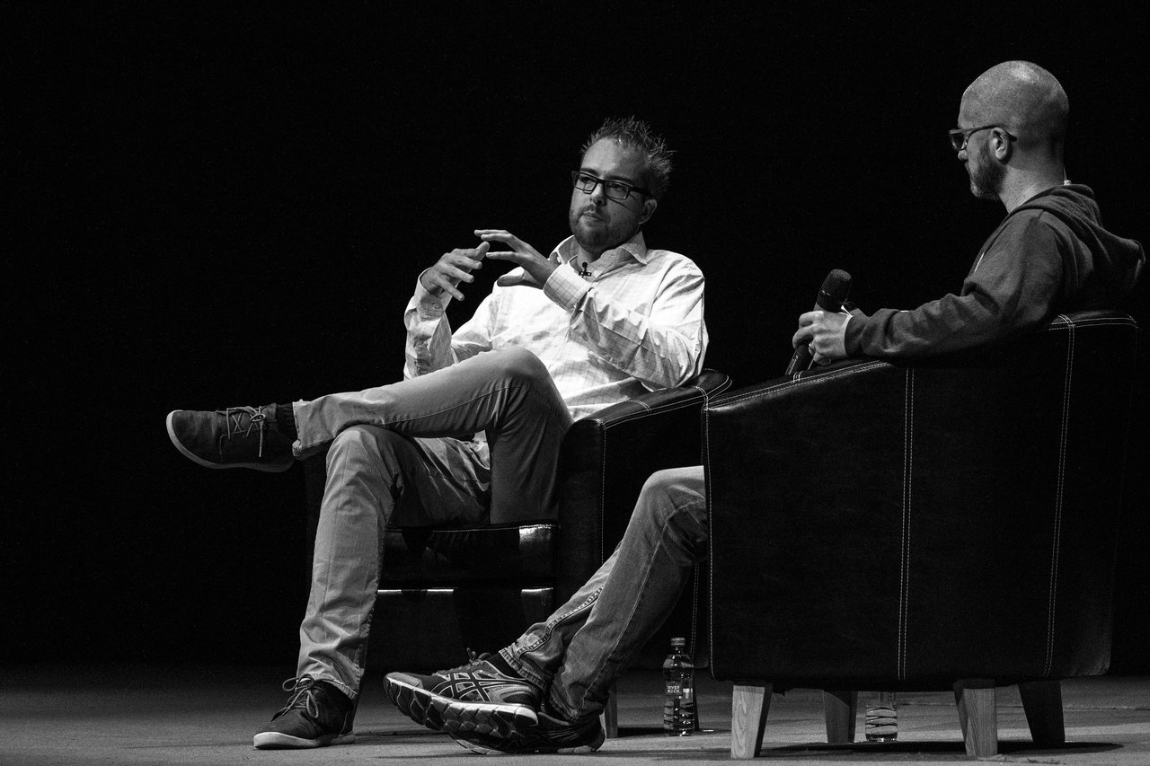 Two people seated on stage in a discussion, one gesturing while speaking and the other holding a microphone.