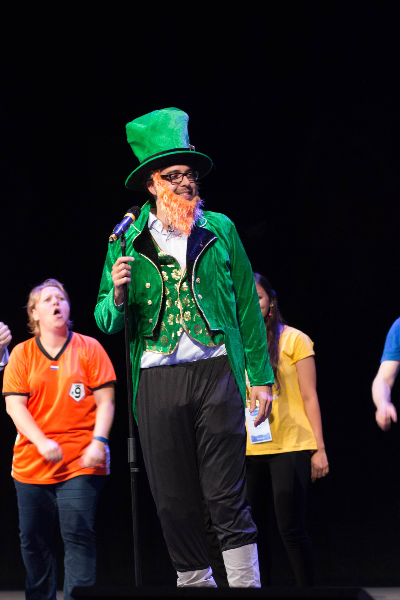 A person in a green leprechaun costume speaks into a microphone on stage, with others reacting behind them.