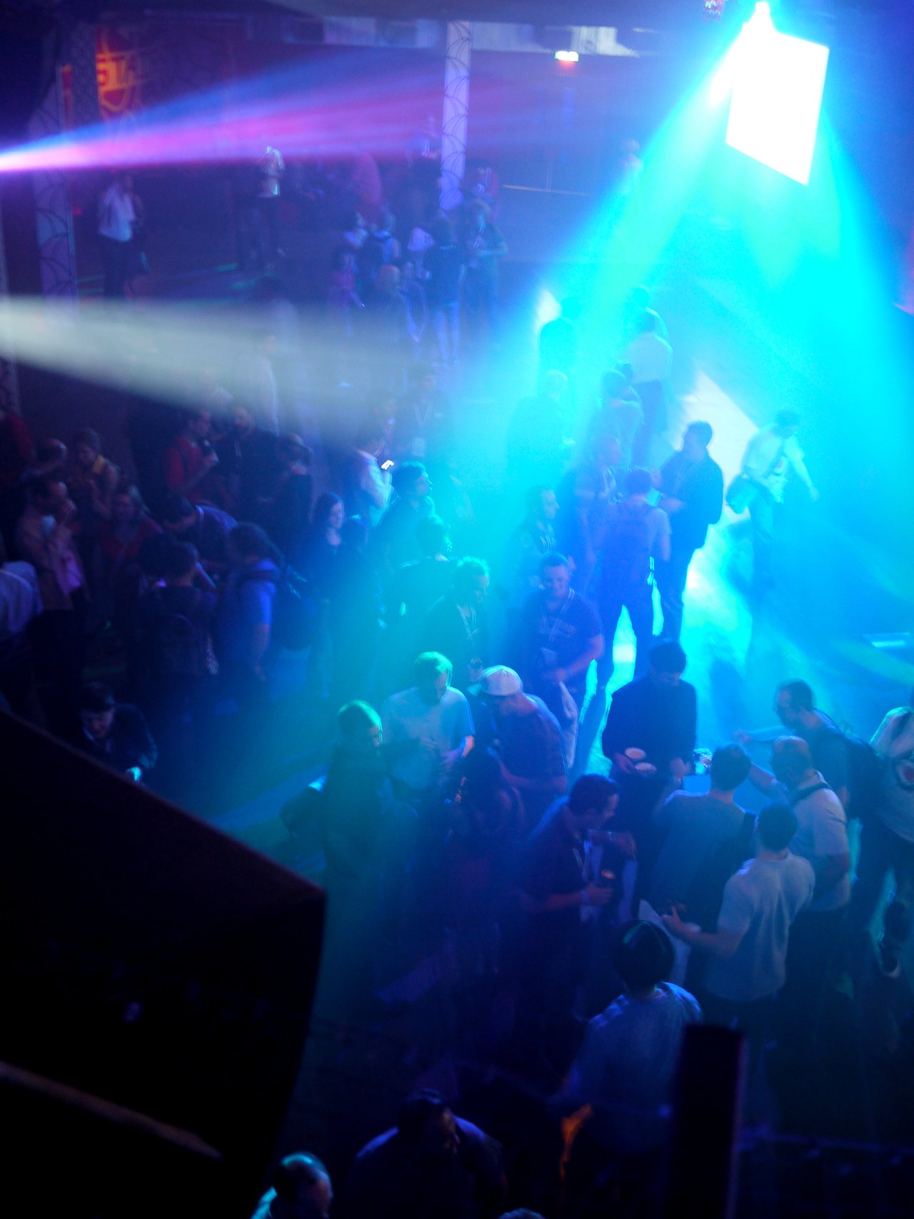 A crowd of people socializing at a party with colorful lights and beams shining across the venue.