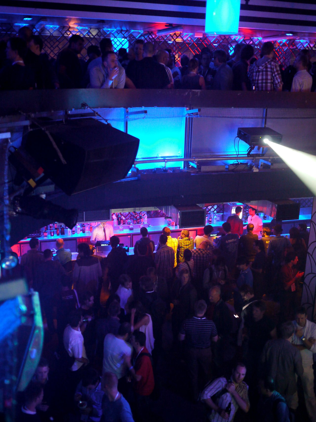 A large crowd gathers at a dimly lit bar with colorful lights during the Acquia-sponsored party at DrupalCon London 2011.