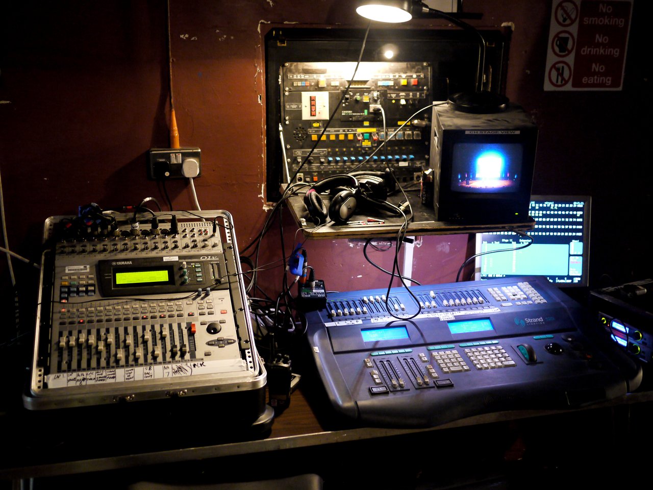 Backstage control setup with audio mixer, lighting console, and monitor screen displaying the keynote stage.