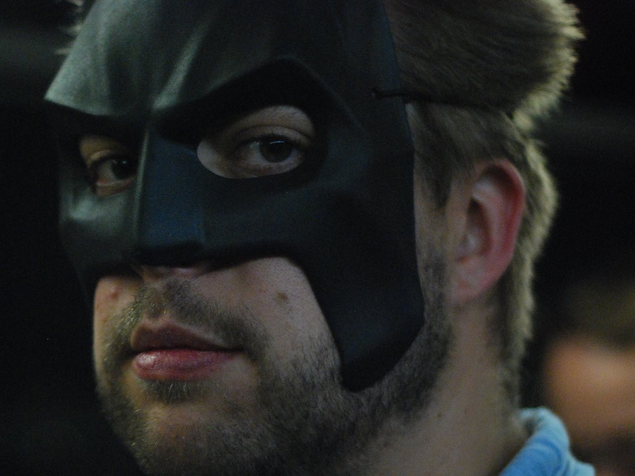 A person wearing a black Batman-style mask looks directly at the camera with a neutral expression.