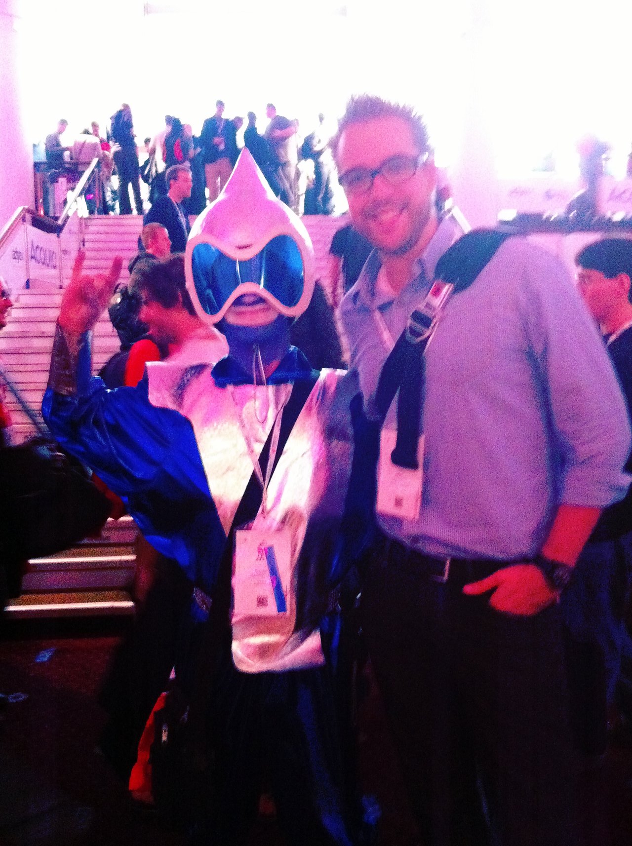 A man in glasses poses with a person dressed in a blue Drupal-themed costume at a conference event.