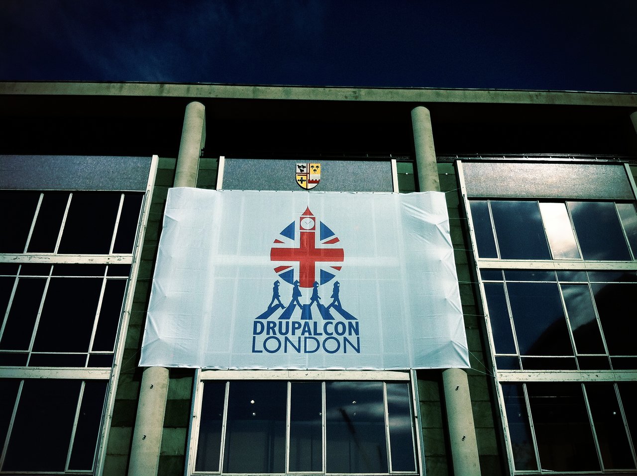 A large DrupalCon London banner hangs on a modern glass building, featuring a Union Jack-inspired design with silhouettes.