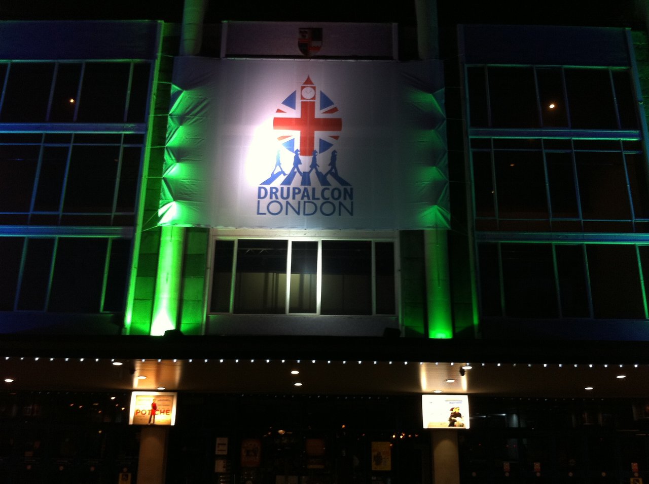 A large DrupalCon London banner is displayed on a building at night, illuminated by green and white lights.