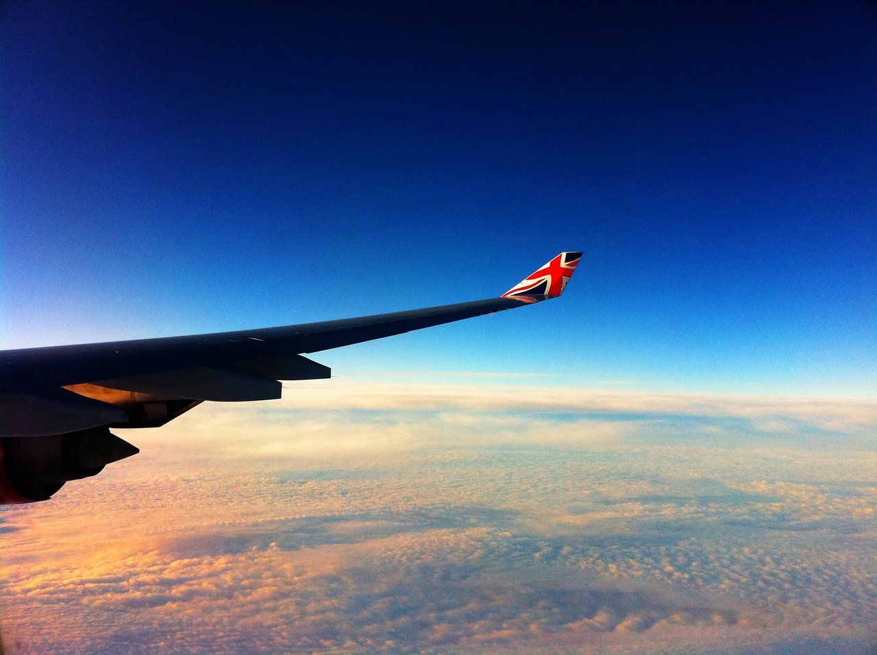 View from an airplane window showing a wing with a Union Jack design, flying above clouds toward London.