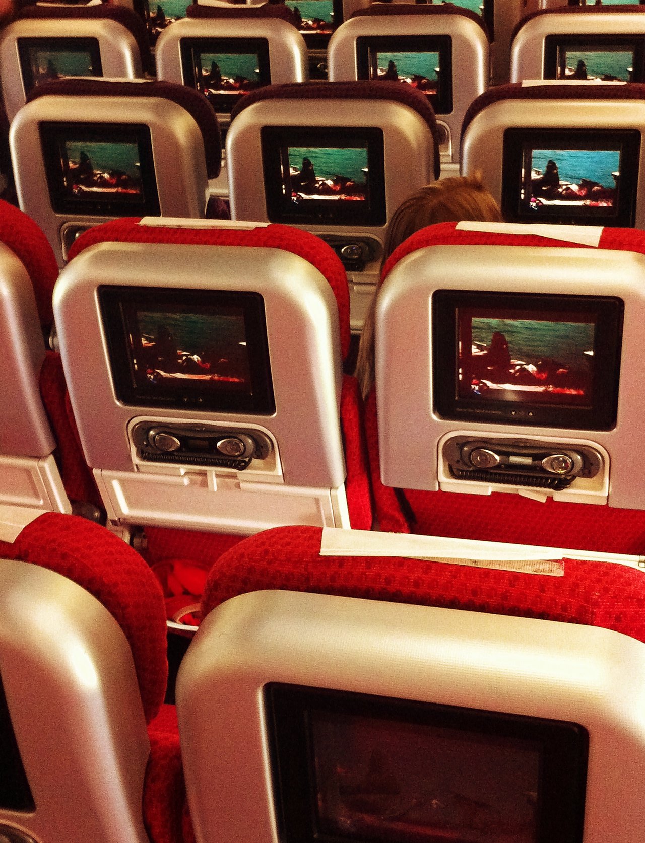 Airplane cabin with red seats and multiple seatback screens displaying the same movie scene during a flight to London.