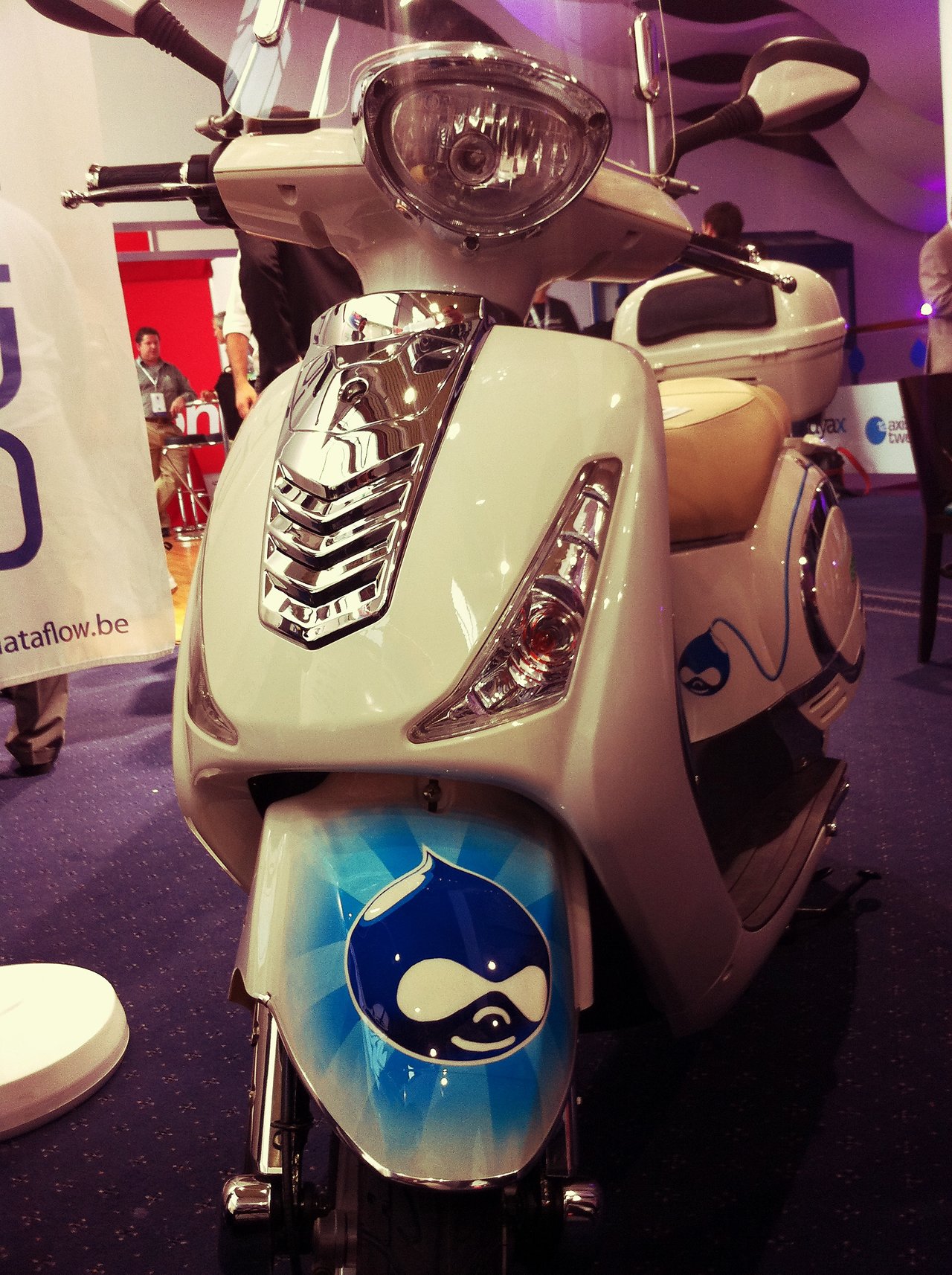 White scooter with a hand-painted Drupal logo on the front fender, displayed indoors at DrupalCon London 2011.