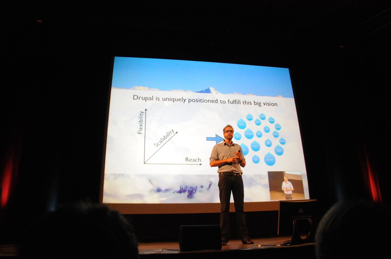 A speaker on stage presents a keynote about Drupal, standing in front of a large screen displaying a slide.