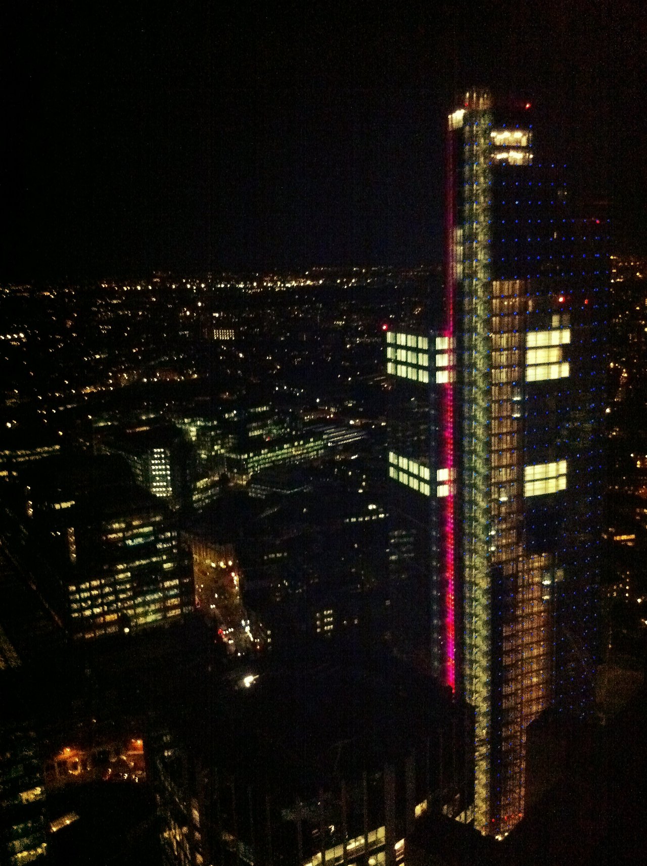 A tall, illuminated skyscraper stands against the night sky, surrounded by city lights and buildings in London.