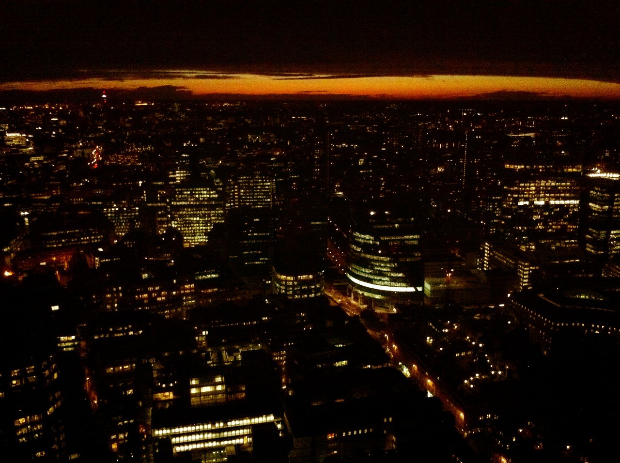 A nighttime aerial view of London with illuminated buildings and streets, and a dark sky with an orange horizon.