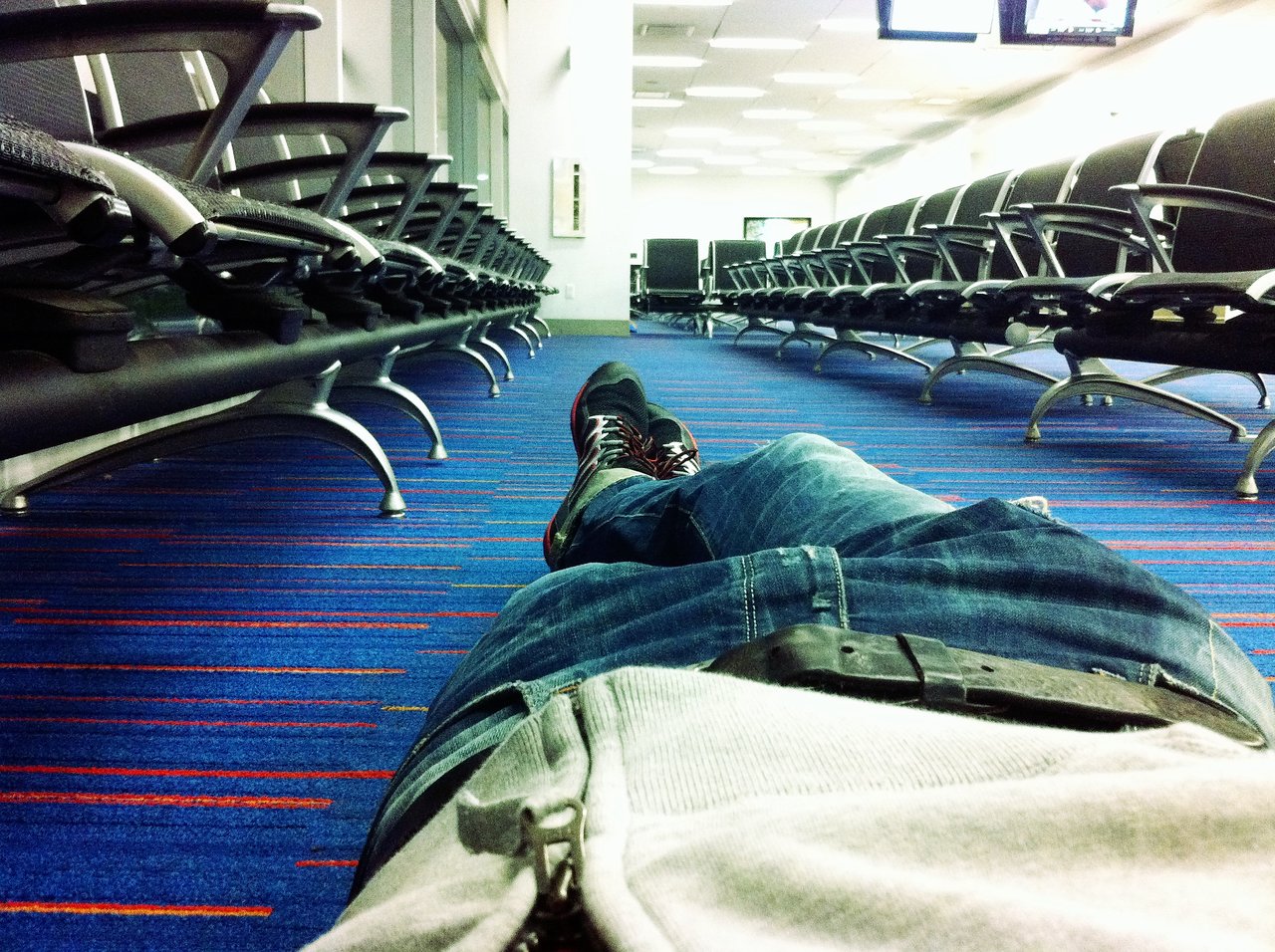 Person lying on the floor in an airport waiting area, wearing jeans and a hoodie, with empty seats nearby.