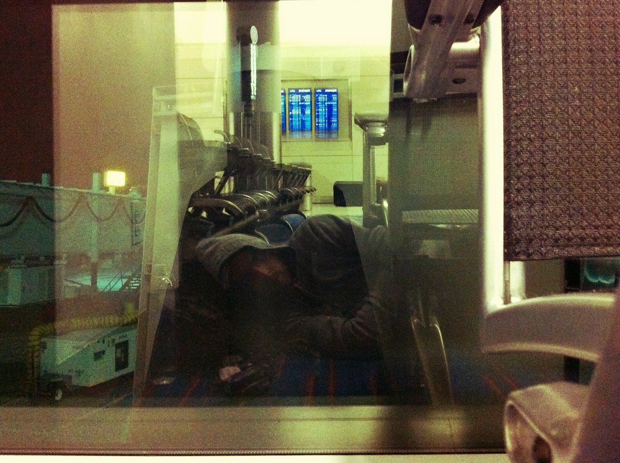 A person in a hoodie is lying down and sleeping on airport seats near a window at JFK Airport.