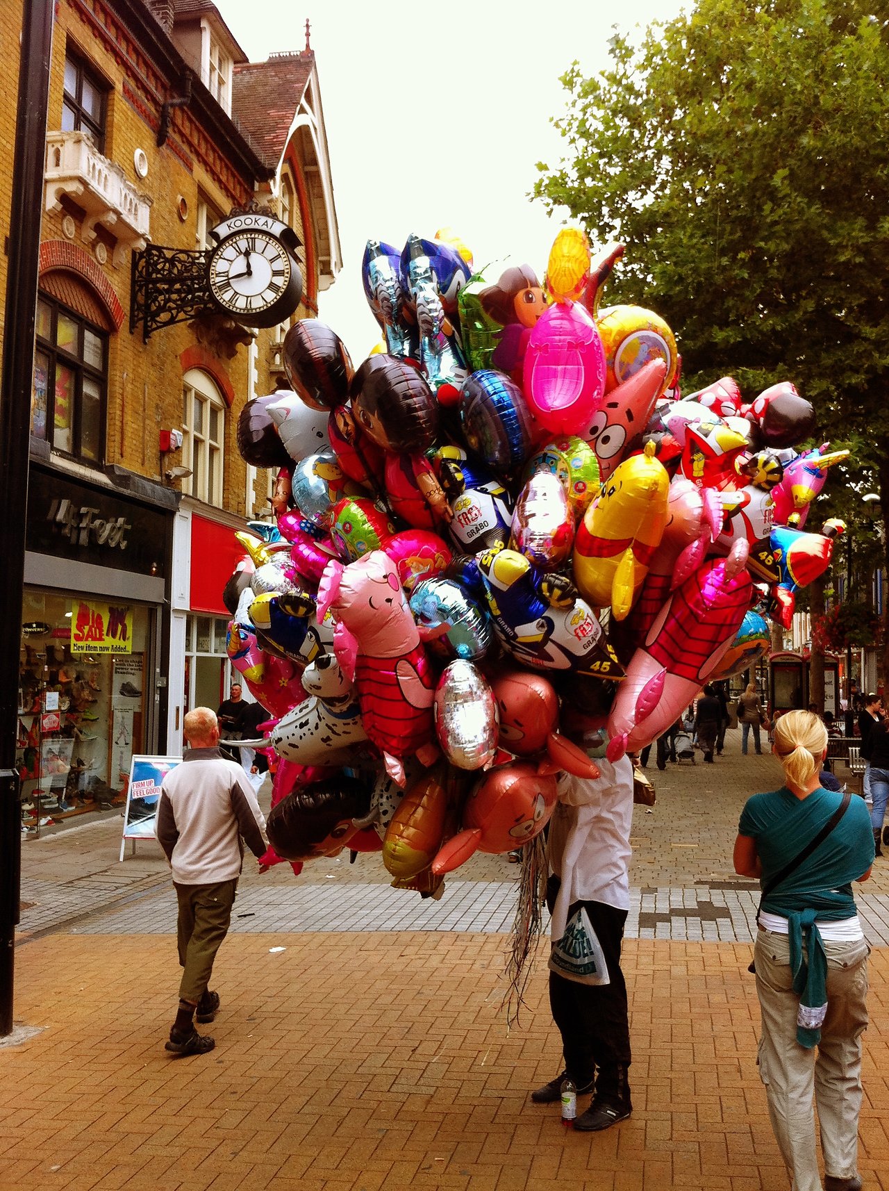 A street vendor holds a large bunch of colorful character balloons while pedestrians walk by in Croydon.