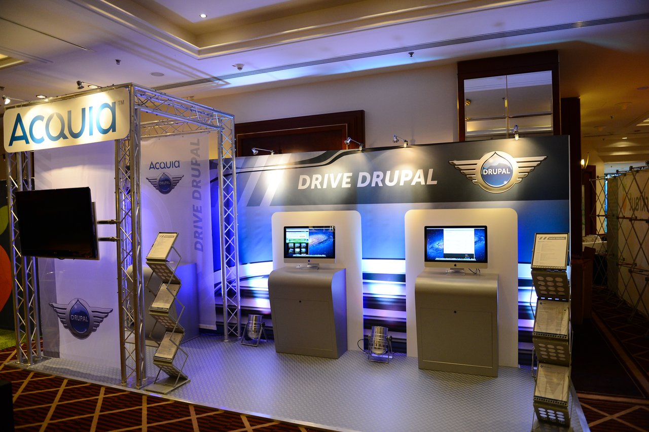 Acquia booth at DrupalCon Munich 2012 featuring two computer stations, promotional materials, and a "Drive Drupal" display.