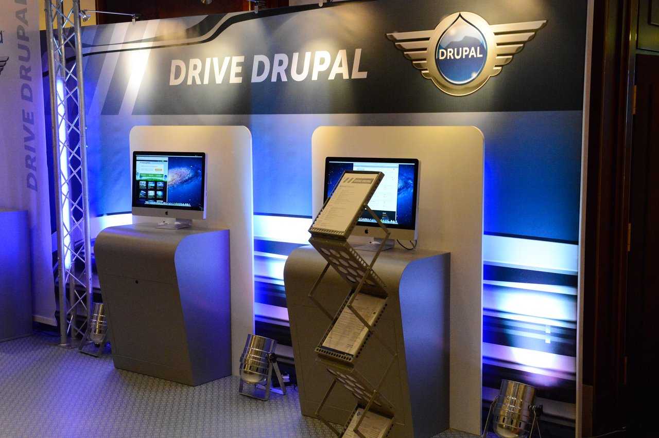 Acquia booth at DrupalCon Munich 2012 with two computer stations, promotional materials, and a "Drive Drupal" backdrop.