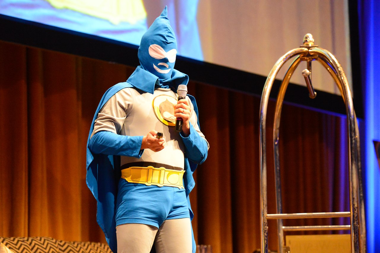 A person in a superhero costume speaks into a microphone on stage during a DrupalCon Munich 2012 keynote.