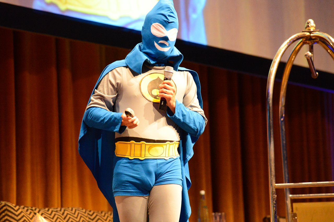 A person in a superhero costume speaks into a microphone on stage during a DrupalCon Munich keynote.