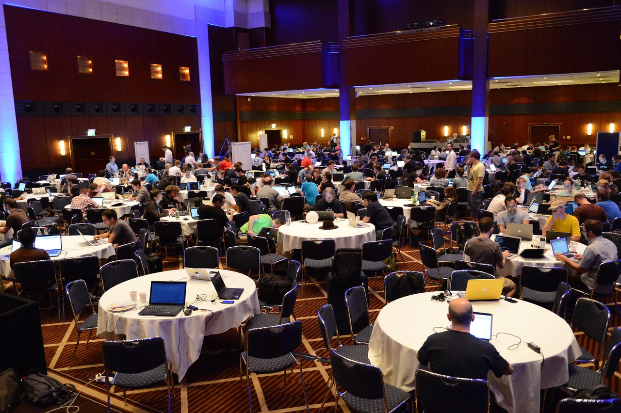 A large group of people working on laptops at round tables during a coding sprint event.
