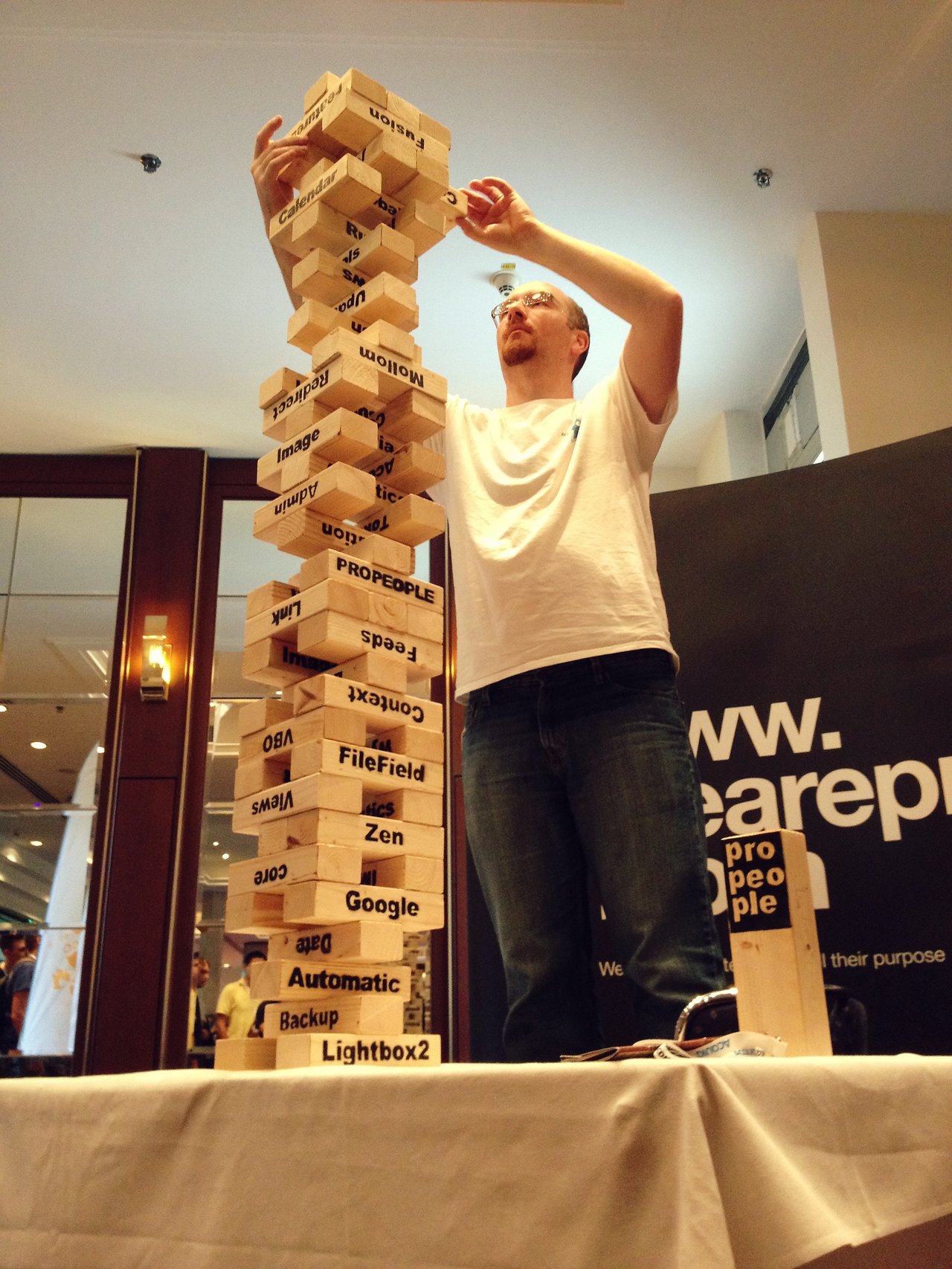 A man carefully places a wooden block on a tall Jenga tower with Drupal-related terms printed on the pieces.