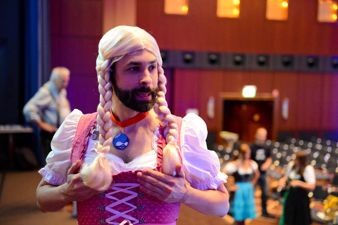 A person in a costume with braided hair and a Drupal logo necklace stands in a conference hall.