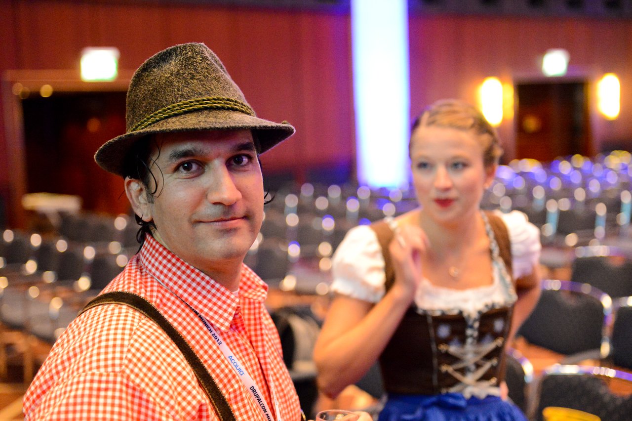 A man in traditional Bavarian clothing looks at the camera, while a woman in similar attire speaks in the background.