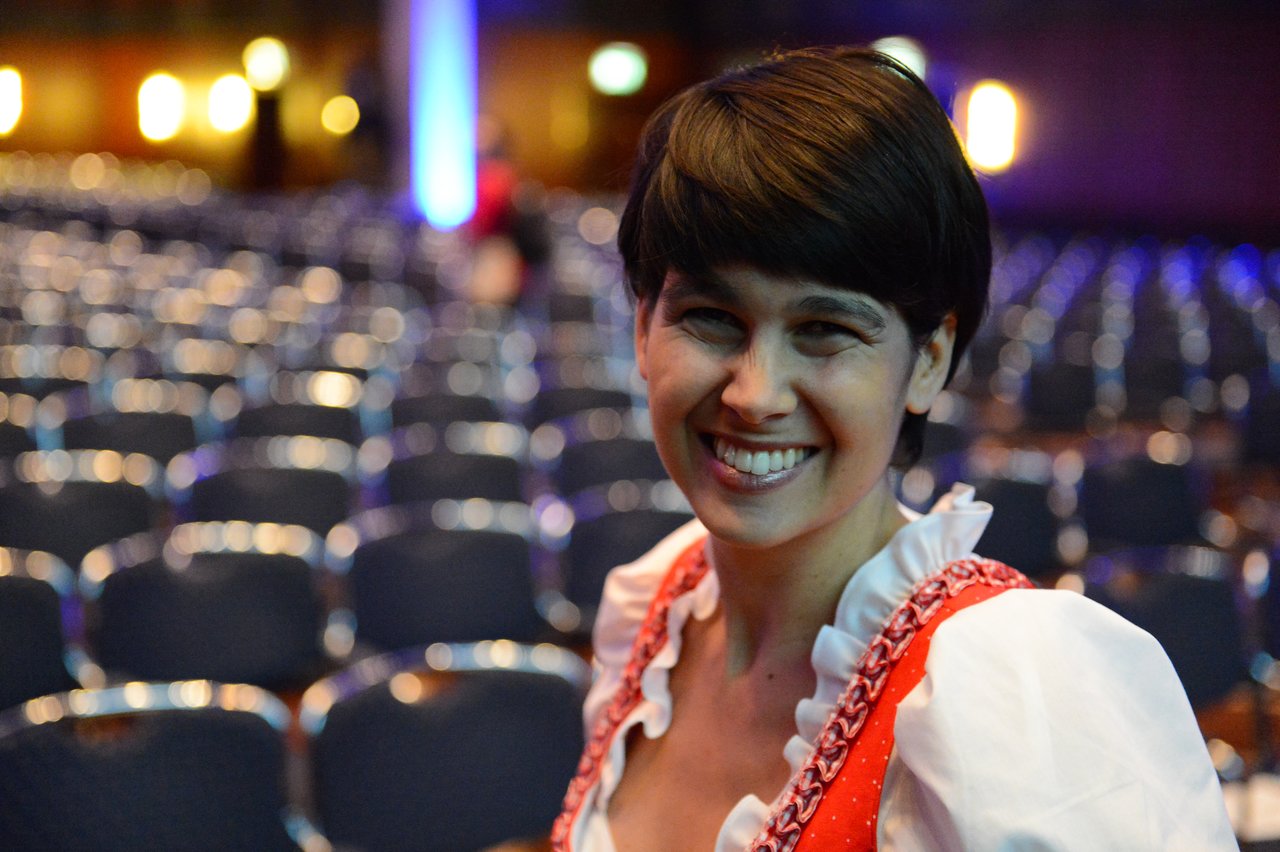 A smiling person in traditional attire stands in front of rows of empty chairs at a conference session.