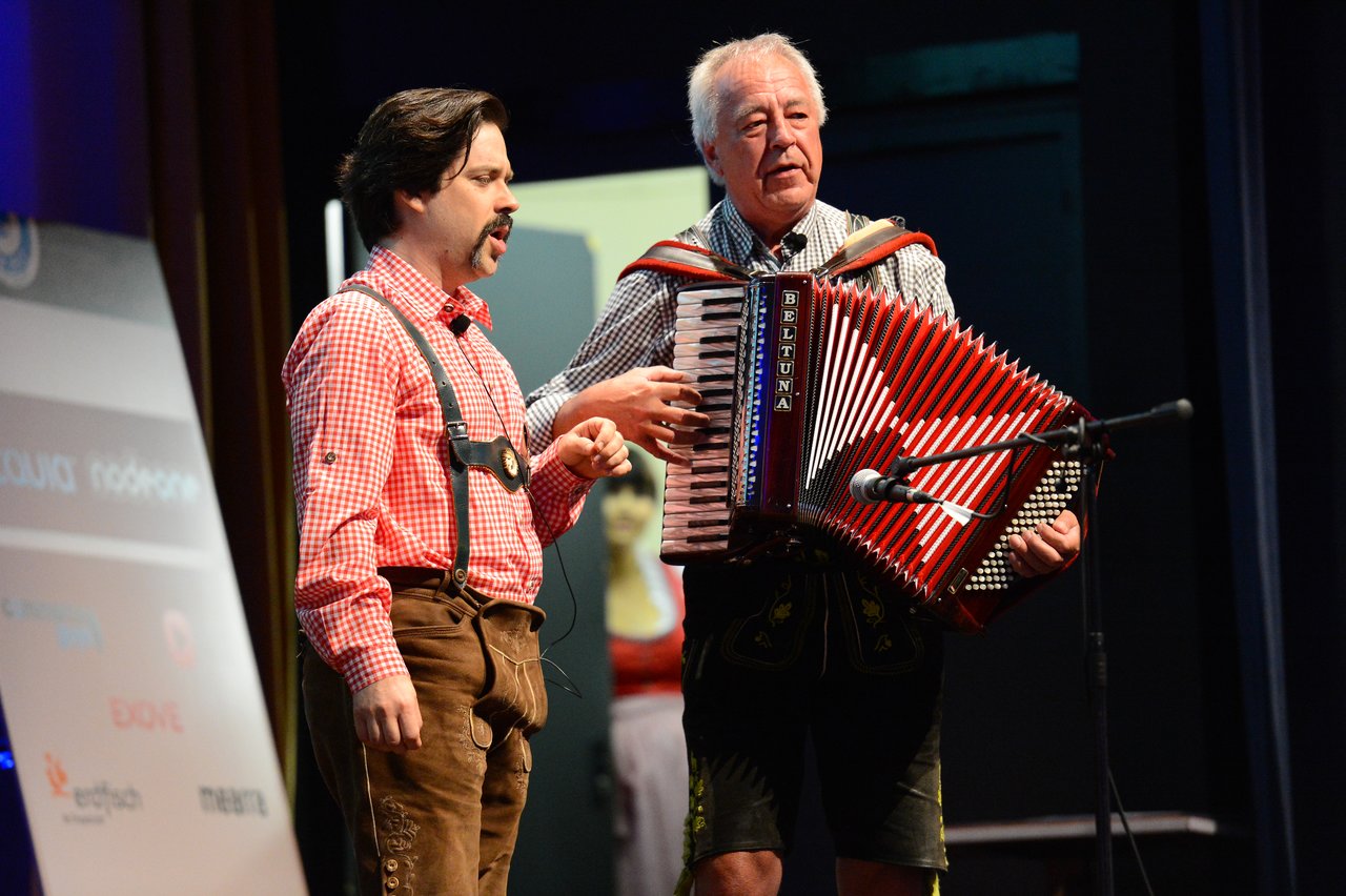 Two men in traditional Bavarian clothing perform on stage, one playing an accordion and the other speaking.
