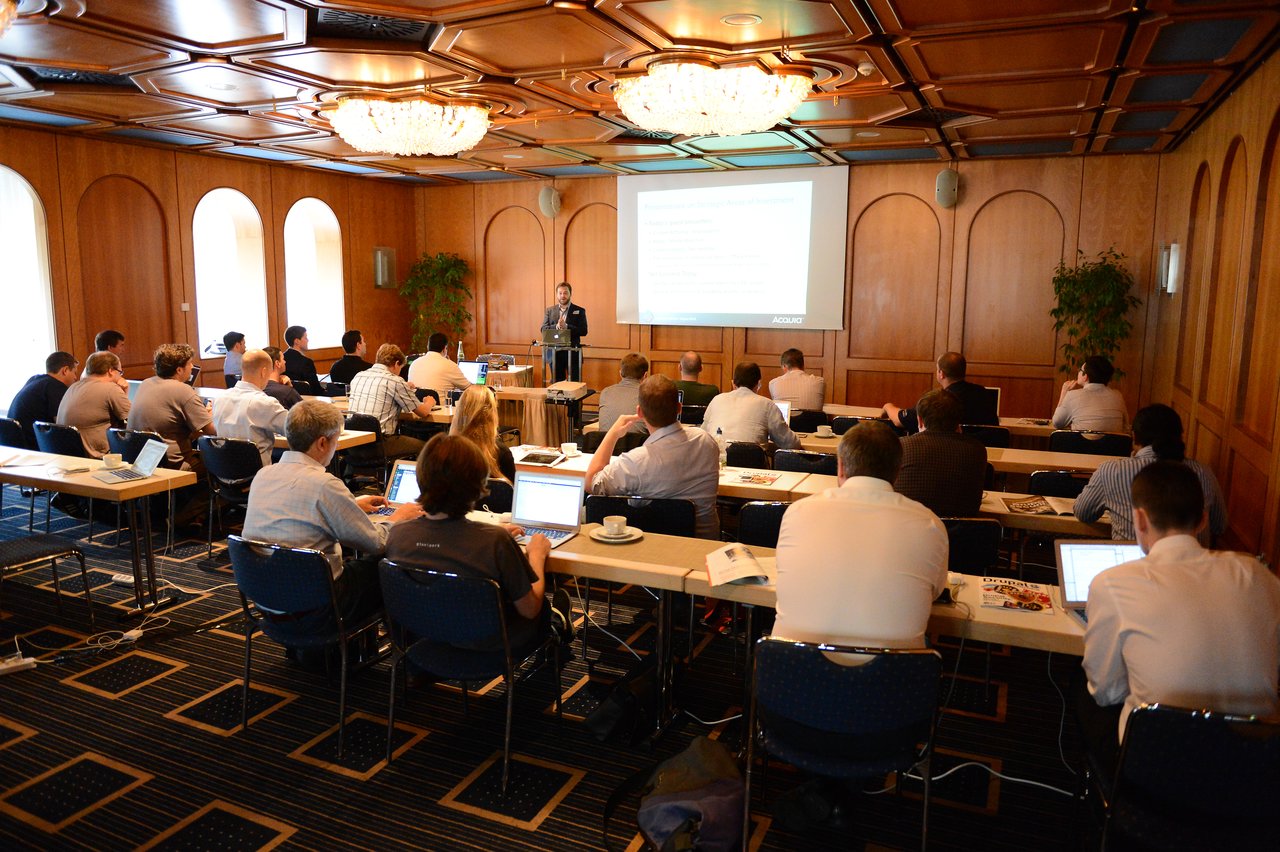 A speaker presents to a room full of attendees using laptops during a large-scale Drupal conference session.