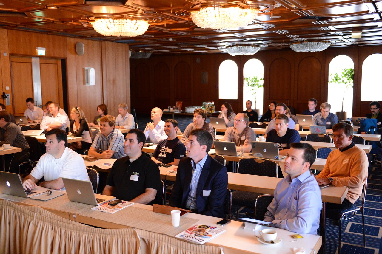 A large group of people attentively listening at a Drupal conference, with many using laptops in a conference room.