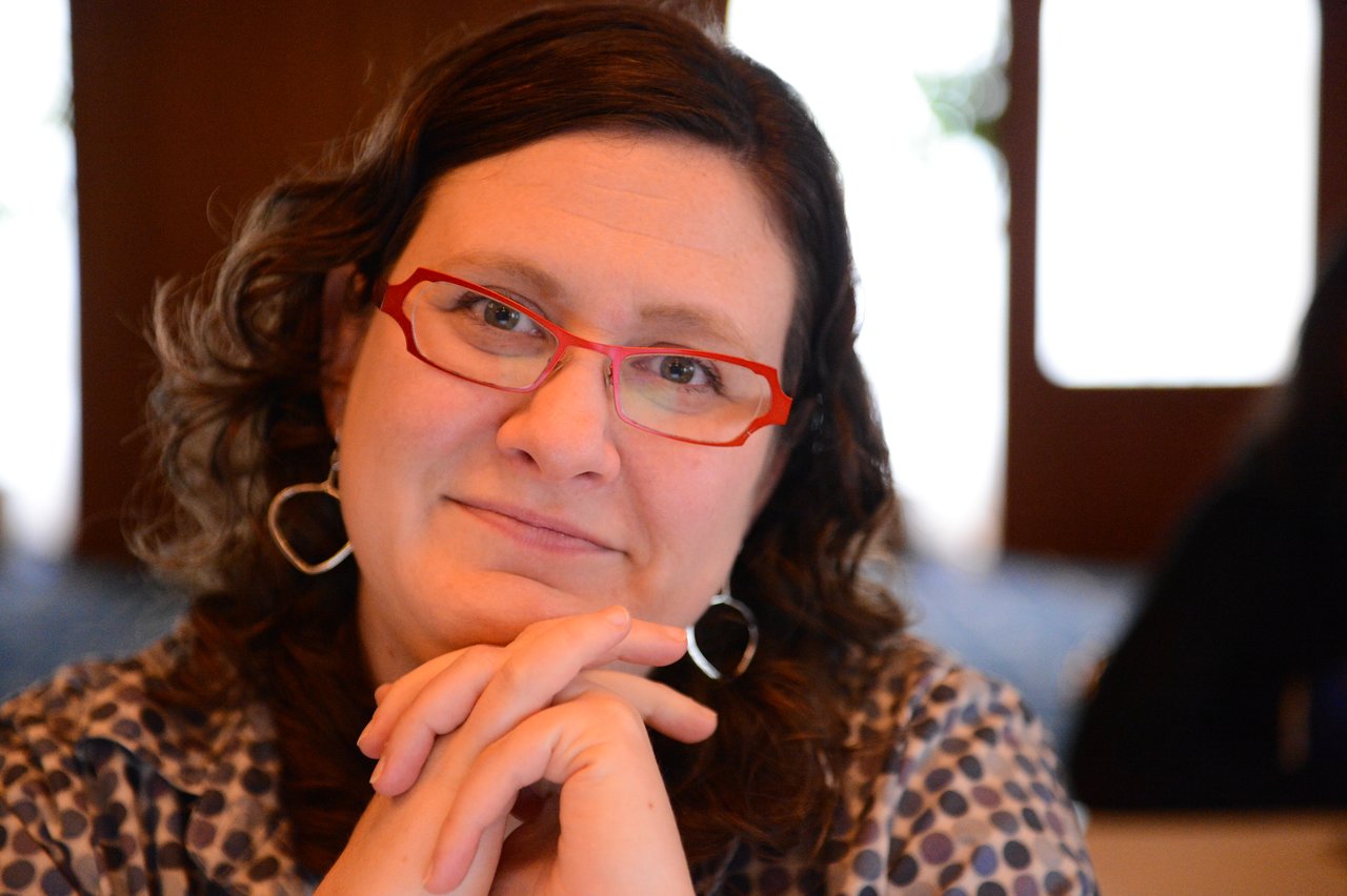 A woman with red glasses and curly hair sits with hands clasped, looking directly at the camera.