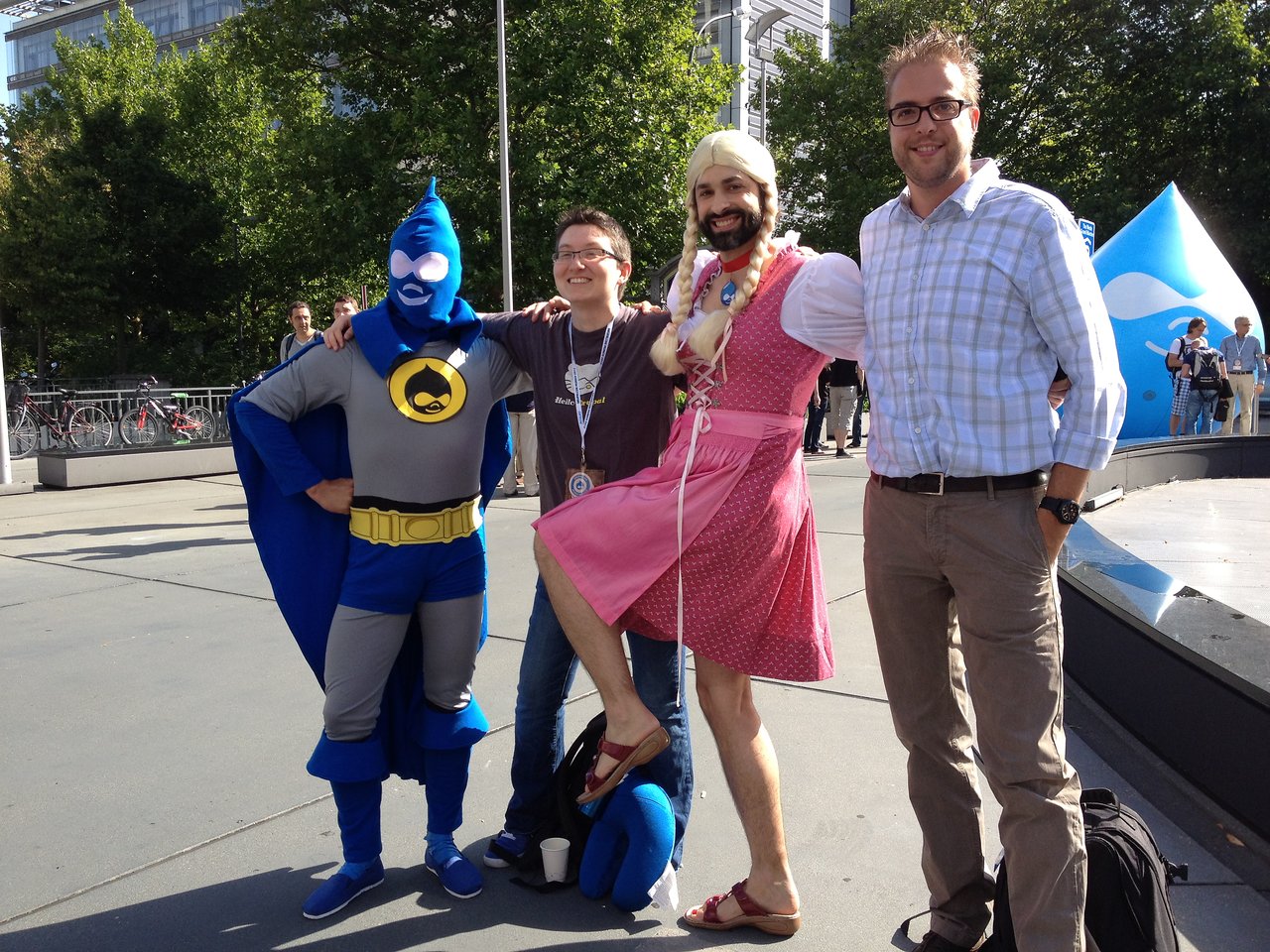 Four people posing together outdoors, including one in a superhero costume and another in a pink dress with a wig.