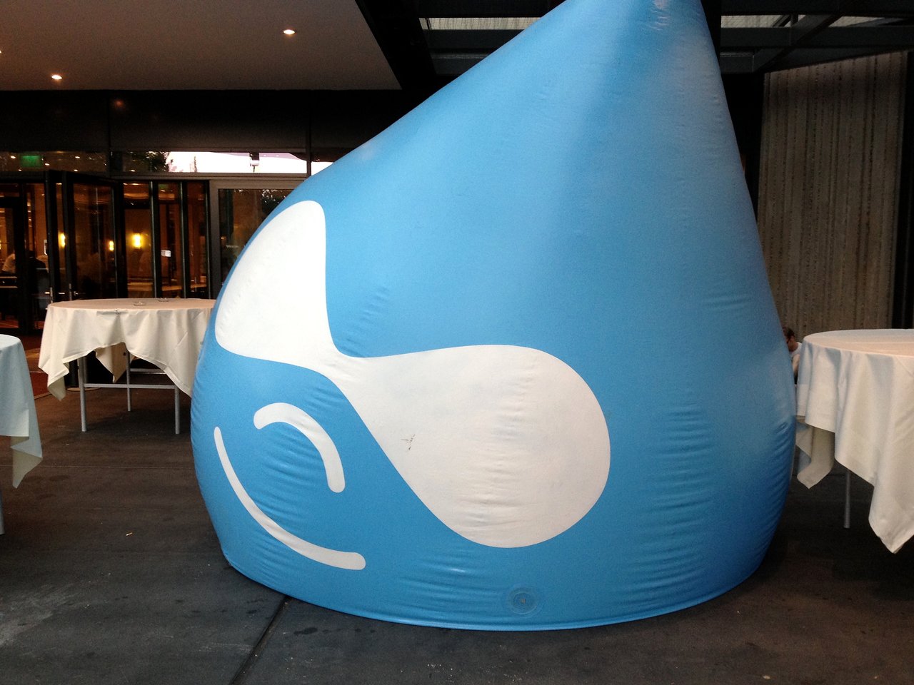 A large, inflatable Druplicon mascot leans to one side near tables at an indoor event space.