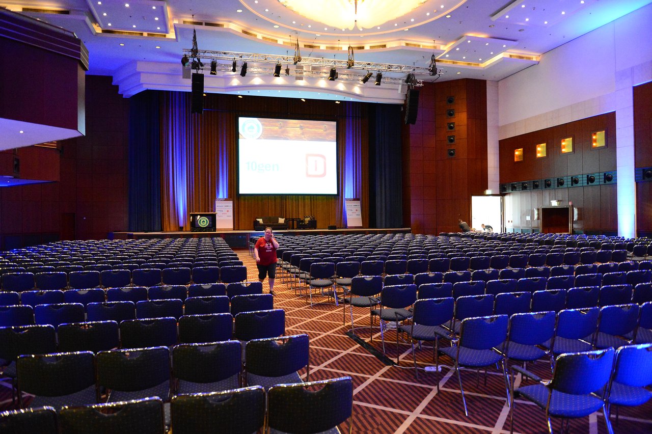 A large conference hall with rows of empty chairs facing a stage with a screen.