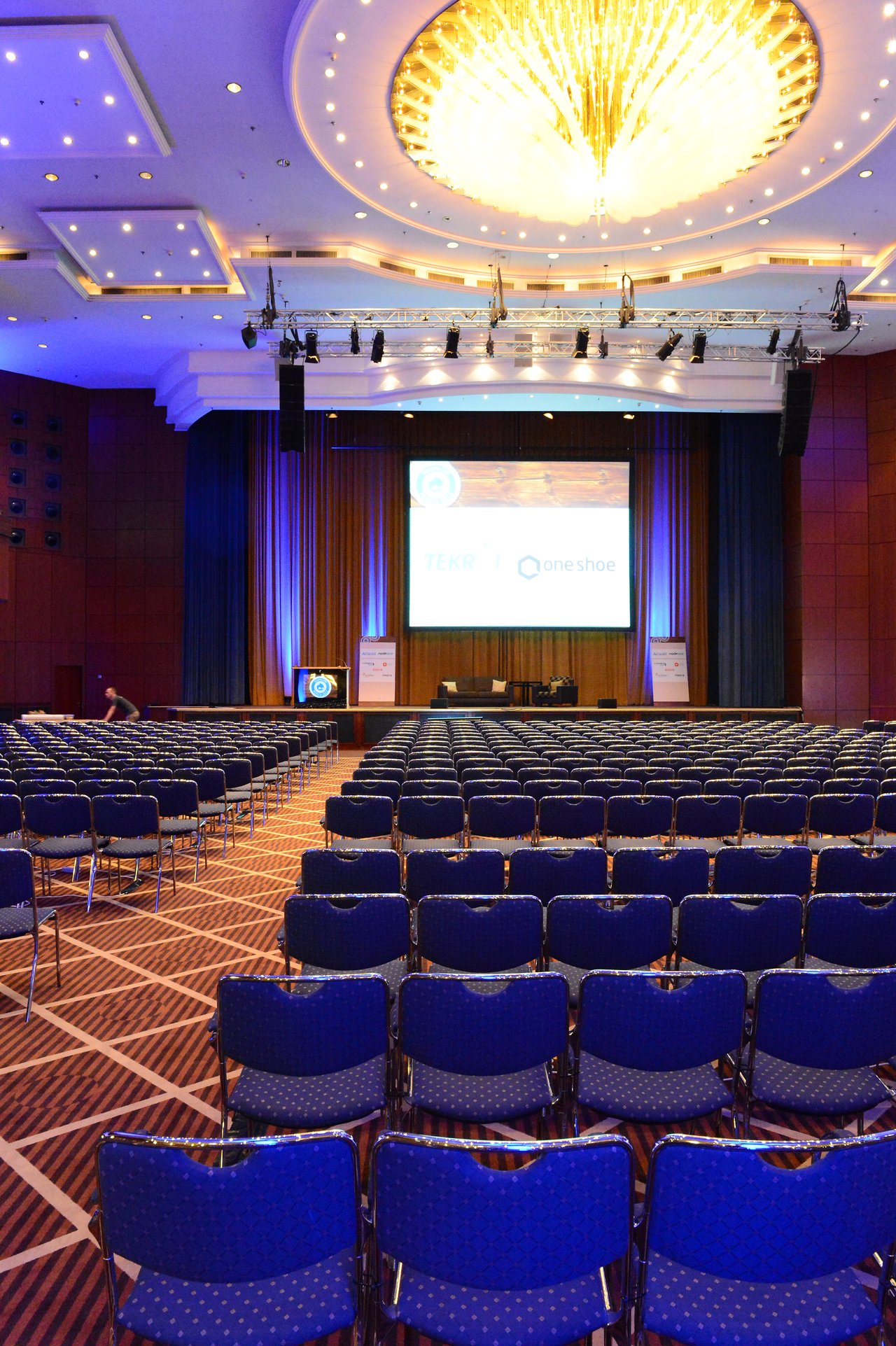 Large conference hall with rows of empty chairs facing a stage with a screen displaying event logos.