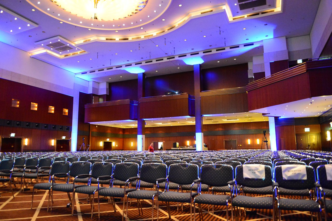 Large conference hall with rows of empty chairs, a few reserved seats, and a person walking near the stage.