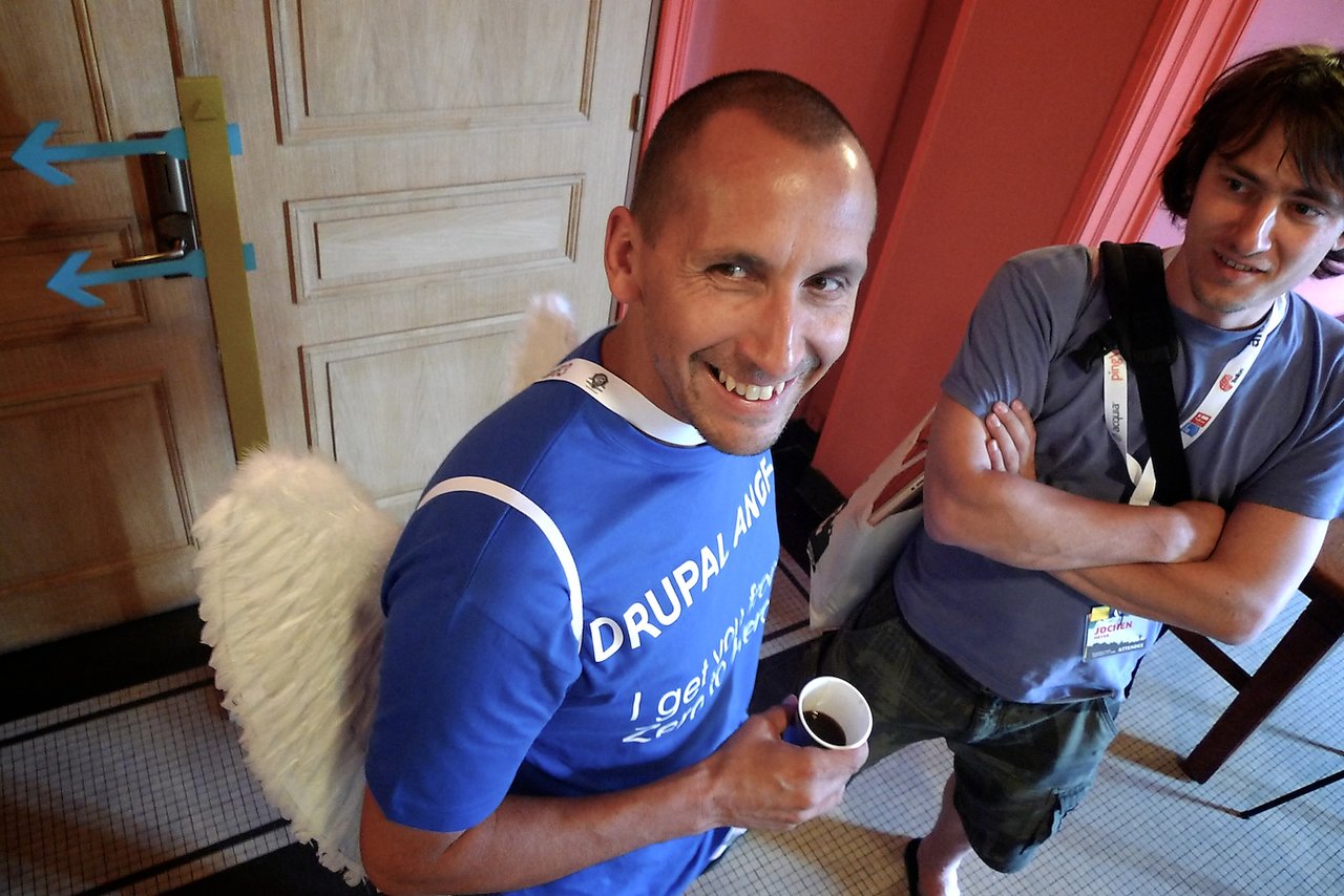 A smiling man in a blue "Drupal Angel" shirt with white wings holds a cup of coffee, standing near another person.