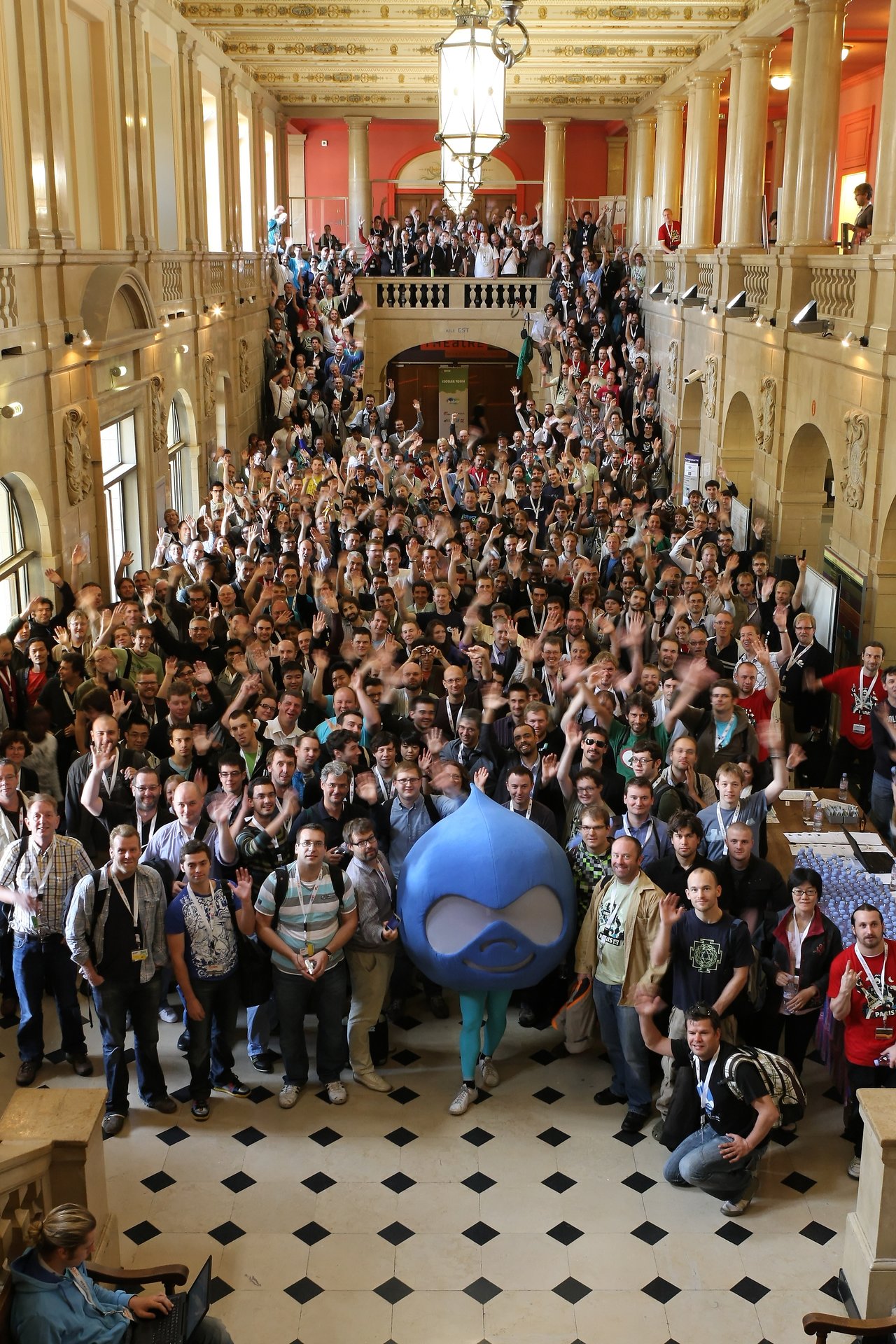 A large group of DrupalCon attendees gathers in a grand hall, waving, with a person in a Drupal mascot costume.