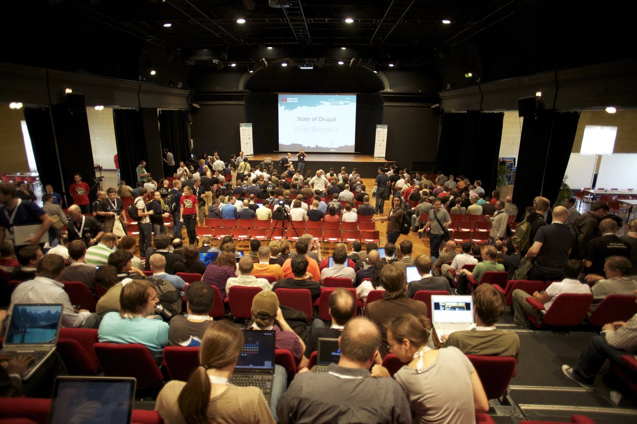 A large audience gathers in a conference hall for a Drupal presentation, with attendees using laptops and conversing.