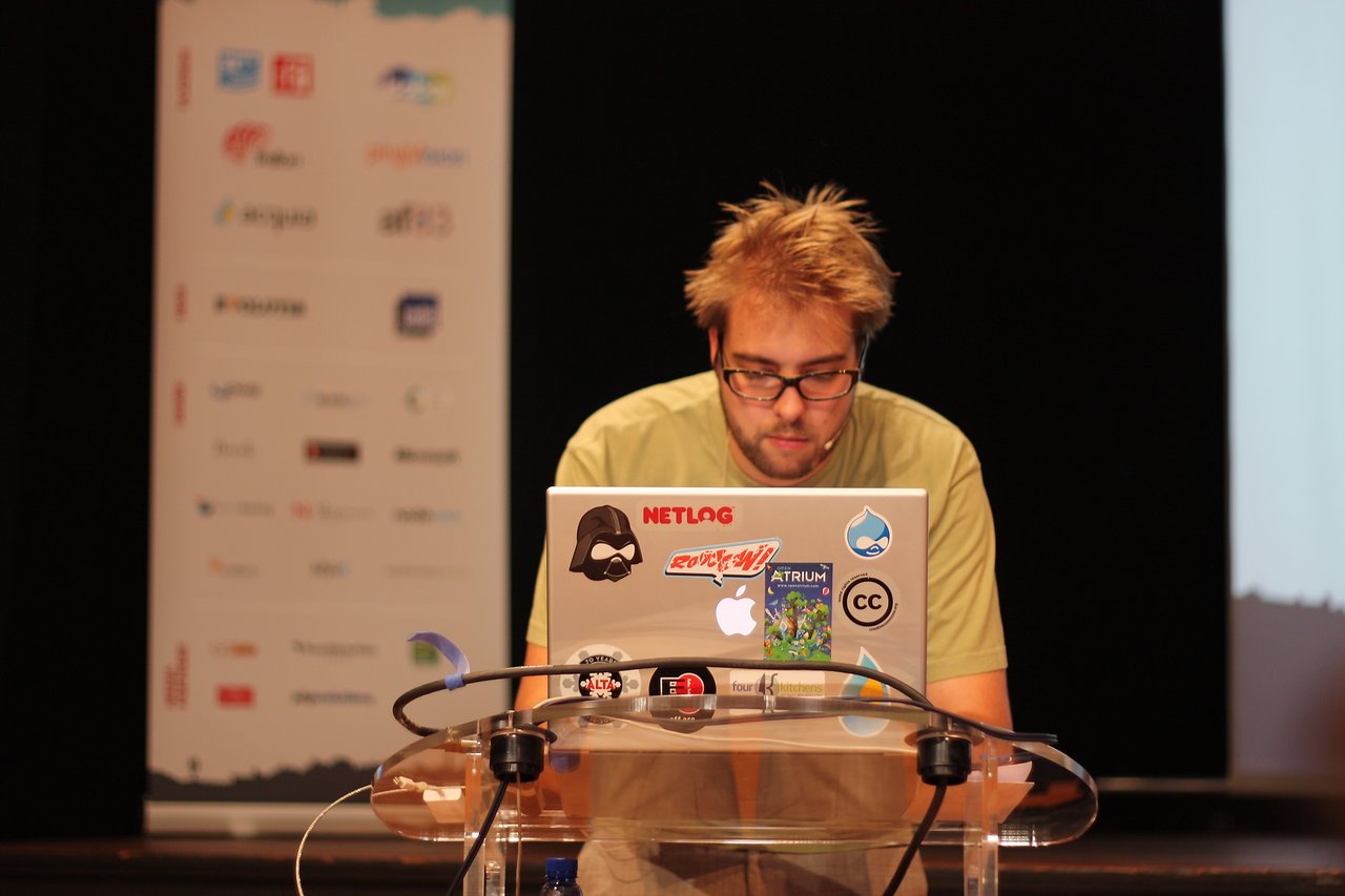 A speaker at DrupalCon Paris 2009 stands at a podium, focused on a laptop covered in stickers.